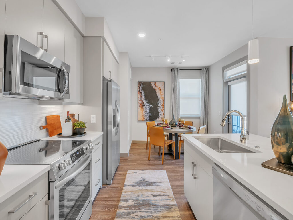 Modern kitchen-dining area with stainless steel appliances, white countertops, and a small round dining table with orange chairs