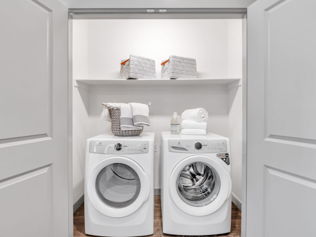 Open closet with side-by-side washer and dryer, shelf above with baskets and cleaning supplies.