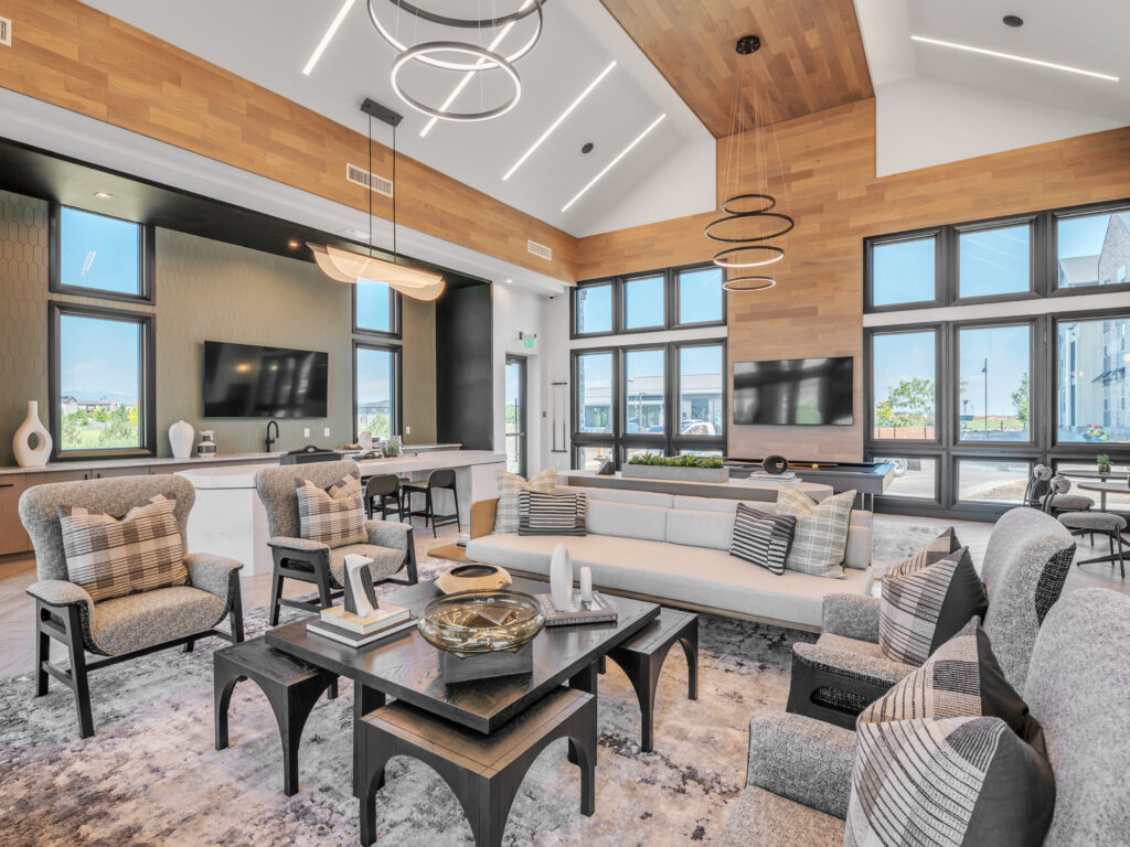 Modern communal lounge with large windows, contemporary furniture, wooden ceiling accents, and two wall-mounted televisions.