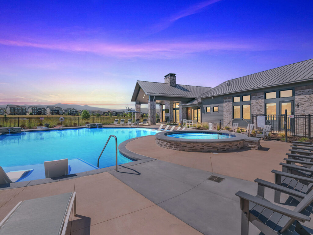 Community pool area at dusk with lounge chairs and a hot tub, surrounded by a modern clubhouse and landscaped grounds.