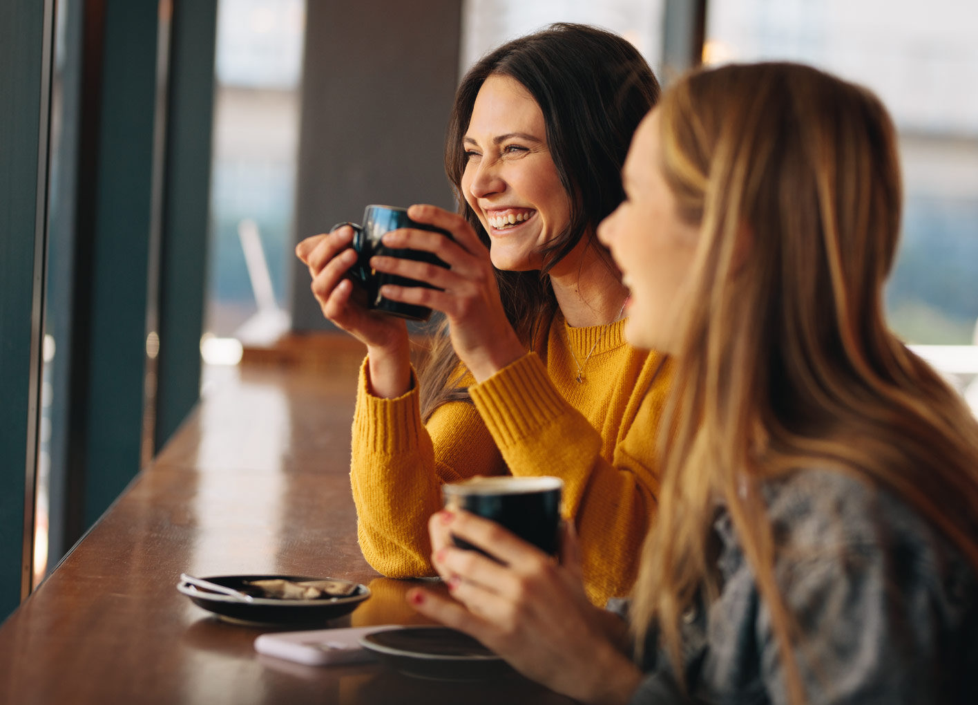 Two women laughing while enjoying coffee inside a cafe.